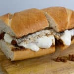 A sandwich on a wooden board, filled with melted cheese and ground meat, with a bread knife nearby and some crumbs on the board.