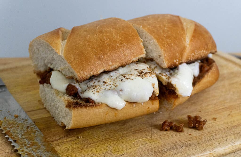 A sandwich on a wooden cutting board, filled with melted cheese and a ground meat mixture, partially sliced with a serrated knife nearby.