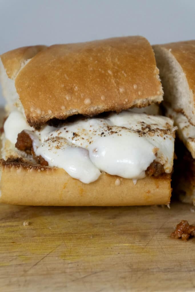 A close-up of a sandwich with melted cheese and a saucy meat filling on a toasted hoagie roll, resting on a wooden surface. The sandwich is cut in half, showing the gooey cheese and hearty filling inside.