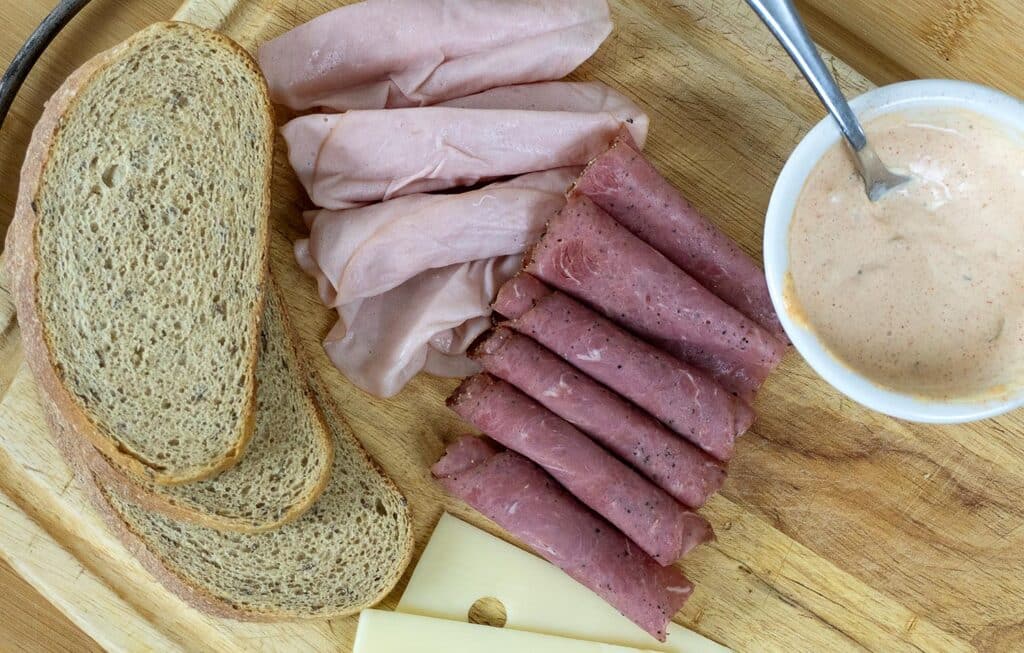 Sliced rye bread, Swiss cheese, rolled pastrami, folded turkey slices, and a bowl of creamy dressing are arranged on a wooden cutting board—everything you need to craft an authentic New Jersey Sloppy Joe.
