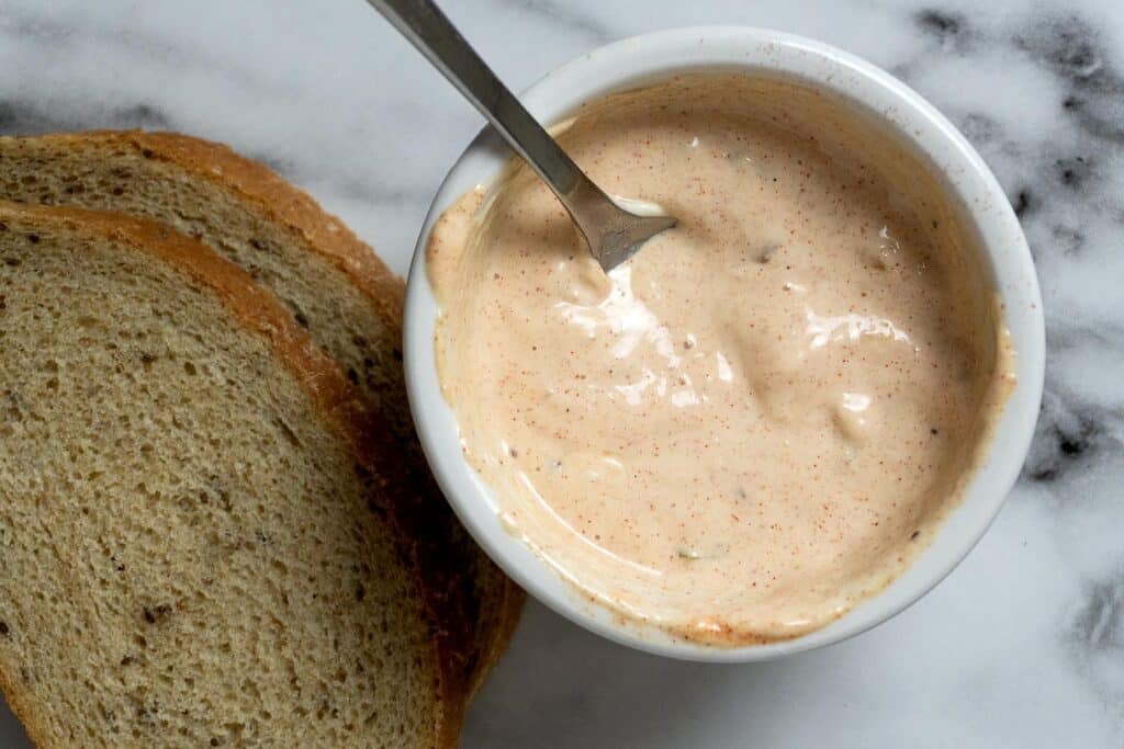 A bowl of creamy, pinkish sauce with a spoon inside sits next to two slices of bread on a marble surface—perfect for making an authentic New Jersey Sloppy Joe sandwich just like you’d find at a classic New Jersey deli.