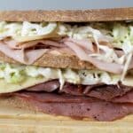 A close-up of a stacked deli sandwich on a wooden board, filled with layers of sliced meats, cheese, and creamy coleslaw between two thick slices of whole grain bread.