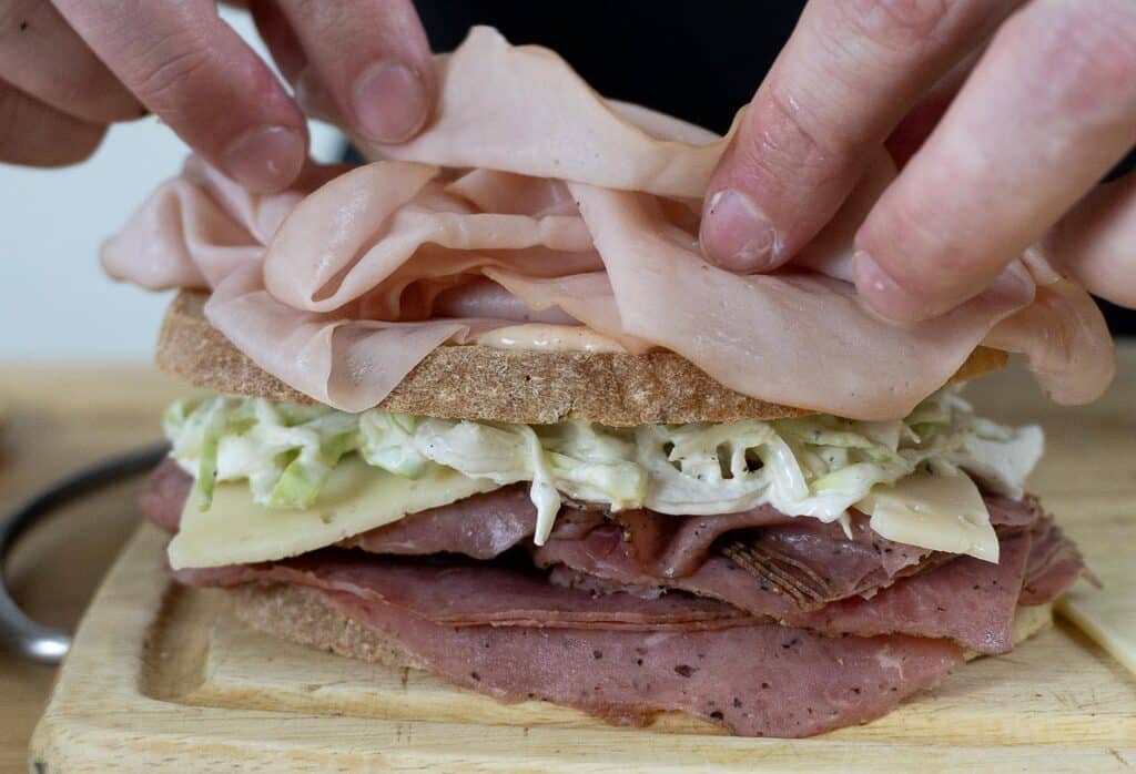 Close-up of hands assembling a New Jersey Sloppy Joe sandwich with layers of sliced deli meat, cheese, and coleslaw on rustic bread, placed on a wooden cutting board.