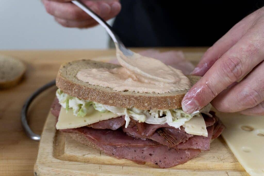 A person spreads sauce on a slice of rye bread to top a classic New Jersey Sloppy Joe sandwich filled with pastrami, Swiss cheese, and coleslaw on a wooden cutting board.