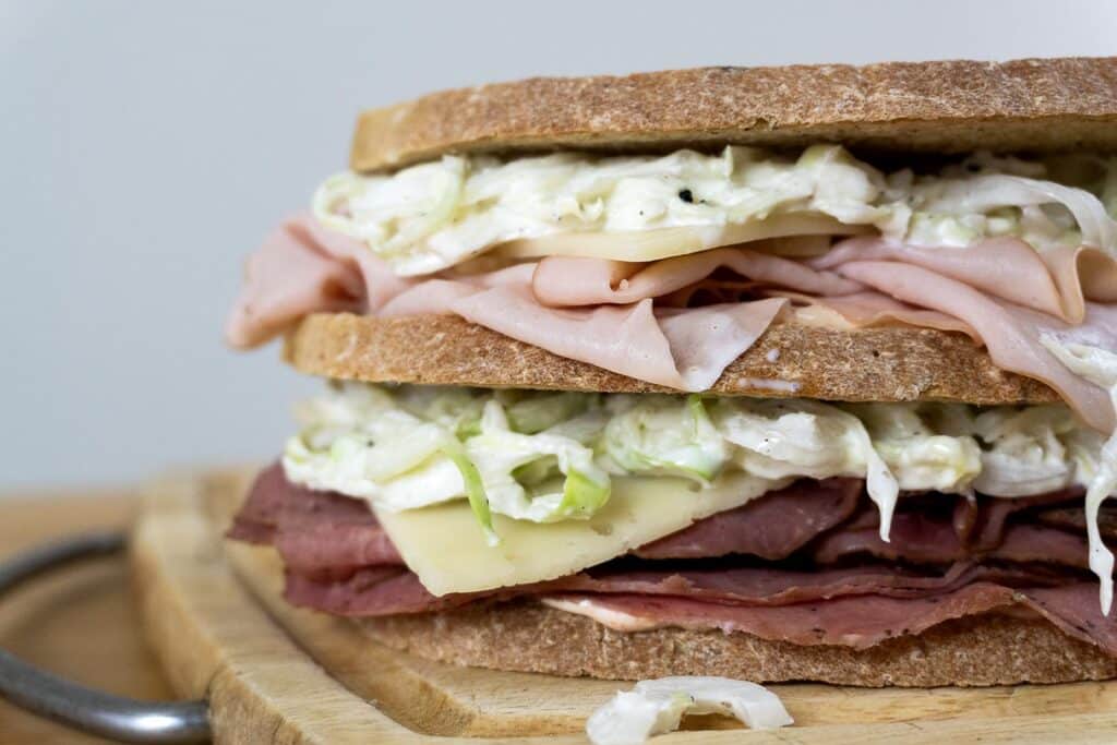 A close-up of a New Jersey Sloppy Joe sandwich on a wooden board, filled with deli meats, cheese slices, and creamy coleslaw between thick slices of bread.