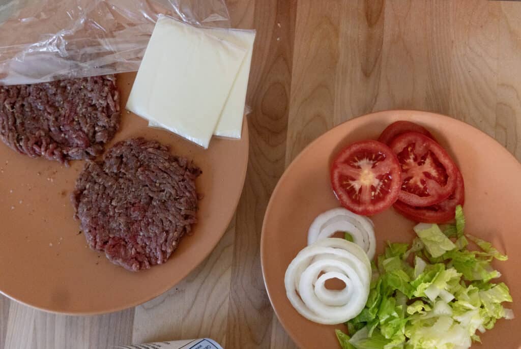 Two uncooked burger patties and two slices of cheese on one plate; on another plate, sliced tomatoes, onion rings, and chopped lettuce are arranged, all set on a wooden surface.