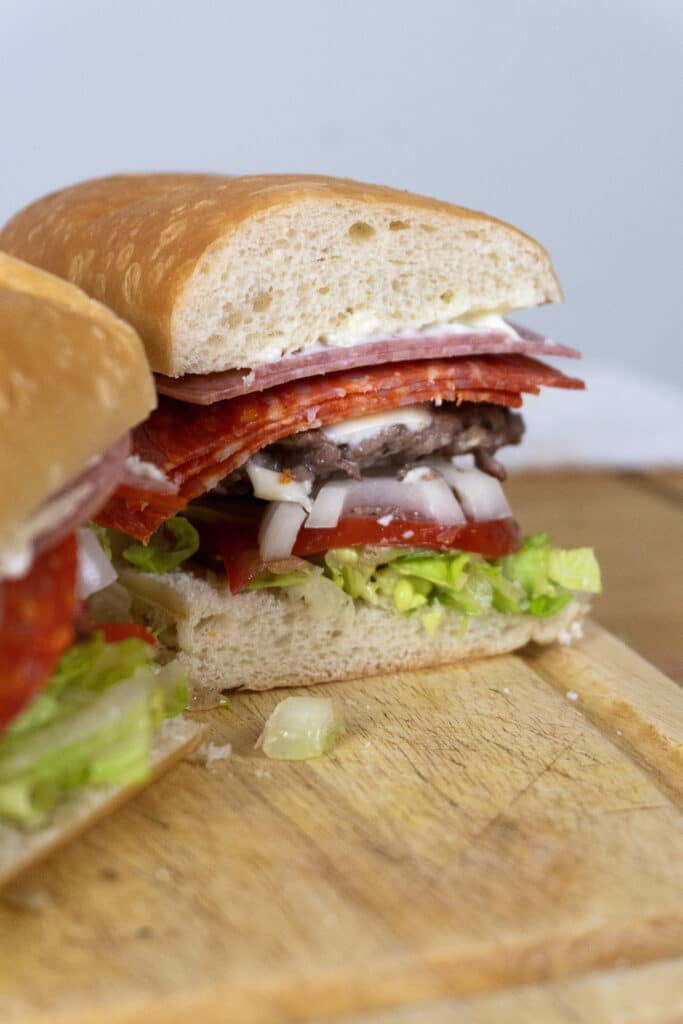 A close-up of a sandwich on a wooden board, filled with layers of lettuce, tomato, chopped onions, sliced deli meats, and mayonnaise on a thick, soft roll.
