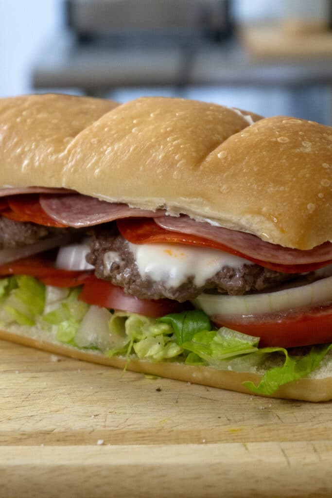 A close-up of a submarine sandwich on a wooden board, filled with lettuce, tomato, onions, hamburger patties, sliced salami, pepperoni, and melted cheese in a soft hoagie roll.
