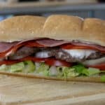 A Roman Burger on a wooden board, filled with lettuce, tomato, onions, sliced beef patty, pepperoni, salami, and mayonnaise in a hoagie roll.