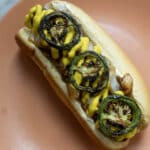 A hot dog in a bun topped with grilled jalapeño slices, sautéed onions, and yellow mustard, served on a peach-colored plate.