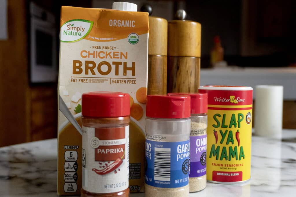 A carton of Simply Nature organic chicken broth, a pair of wooden salt and pepper grinders, and spice containers for paprika, garlic powder, onion powder, and Slap Ya Mama Cajun seasoning on a kitchen counter.
