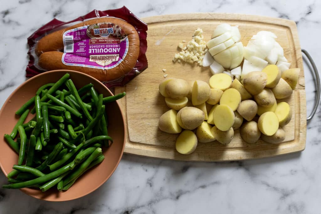 A cutting board on a marble surface with chopped onions, minced garlic, and halved baby potatoes. Next to it are green beans in a bowl and a package of andouille sausage.
