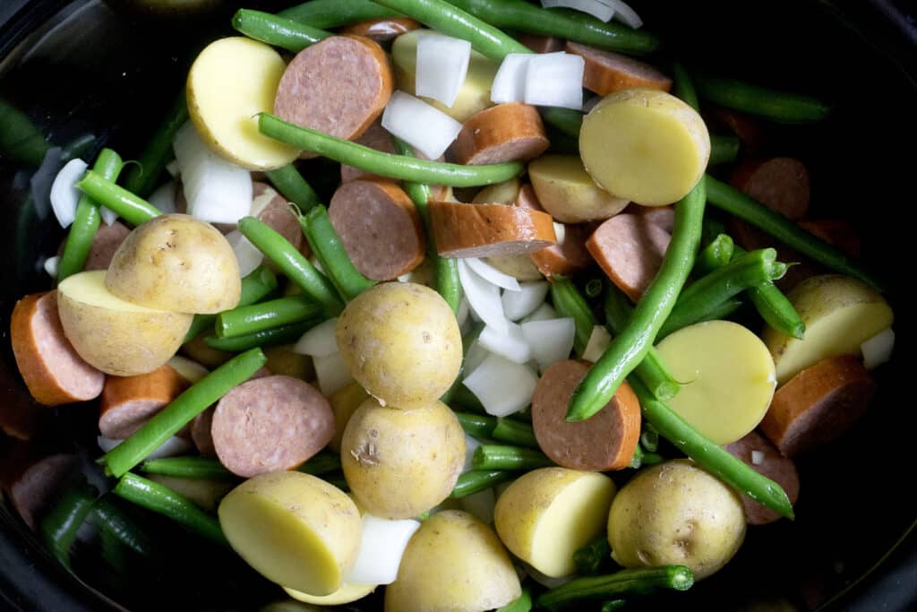 Sliced sausage, halved small potatoes, green beans, and chopped onions are mixed together in a black slow cooker, ready to be cooked.