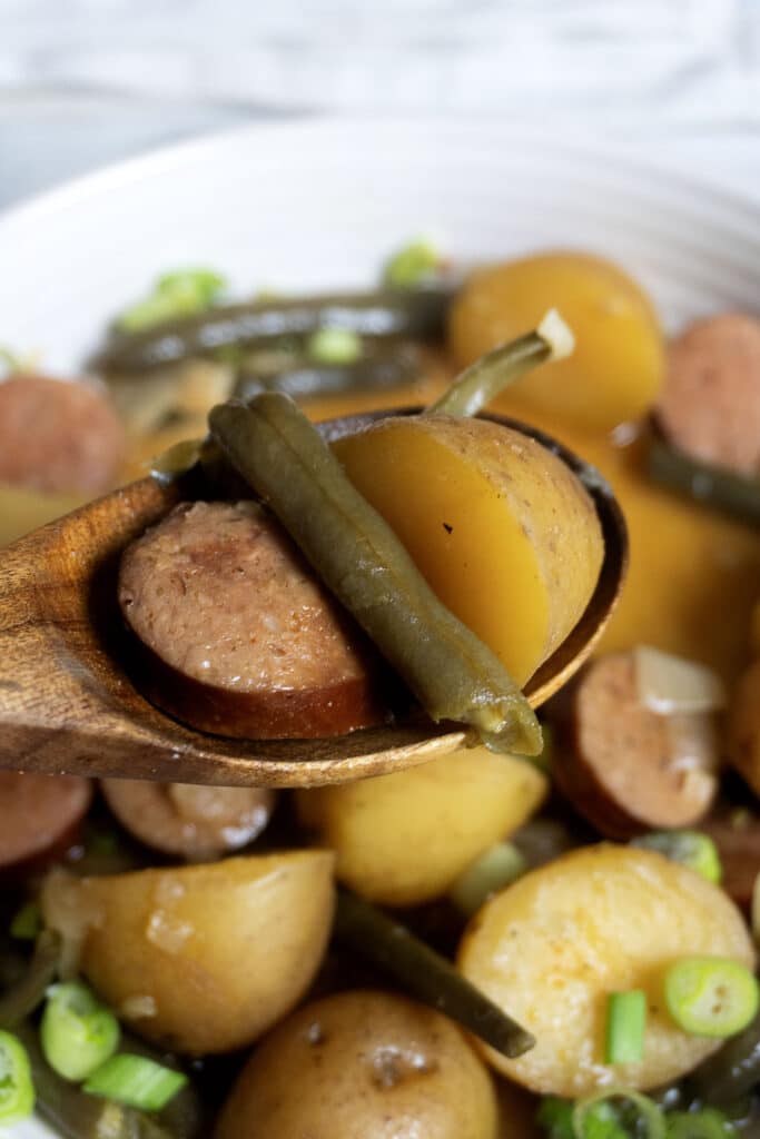 A wooden spoon holds sliced sausage, potatoes, and green beans above a bowl filled with more of the same ingredients in a savory dish.