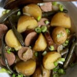 A bowl filled with sliced sausage, halved yellow potatoes, green beans, and chopped green onions, all lightly seasoned and mixed together. A wooden spoon rests on the edge of the bowl.
