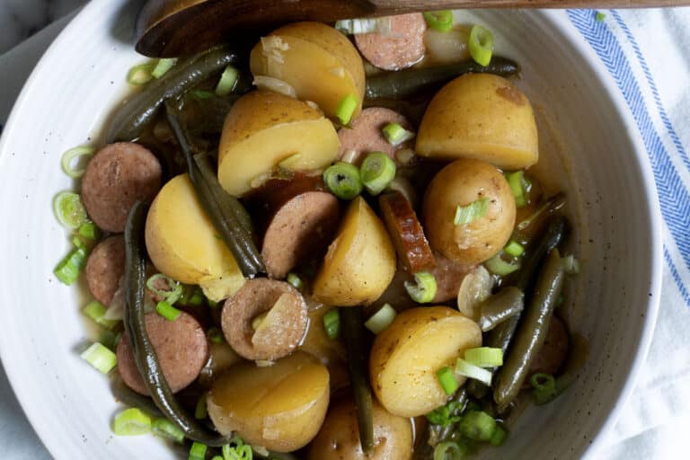 A bowl filled with sliced sausage, halved yellow potatoes, green beans, and chopped green onions, all lightly seasoned and mixed together. A wooden spoon rests on the edge of the bowl.