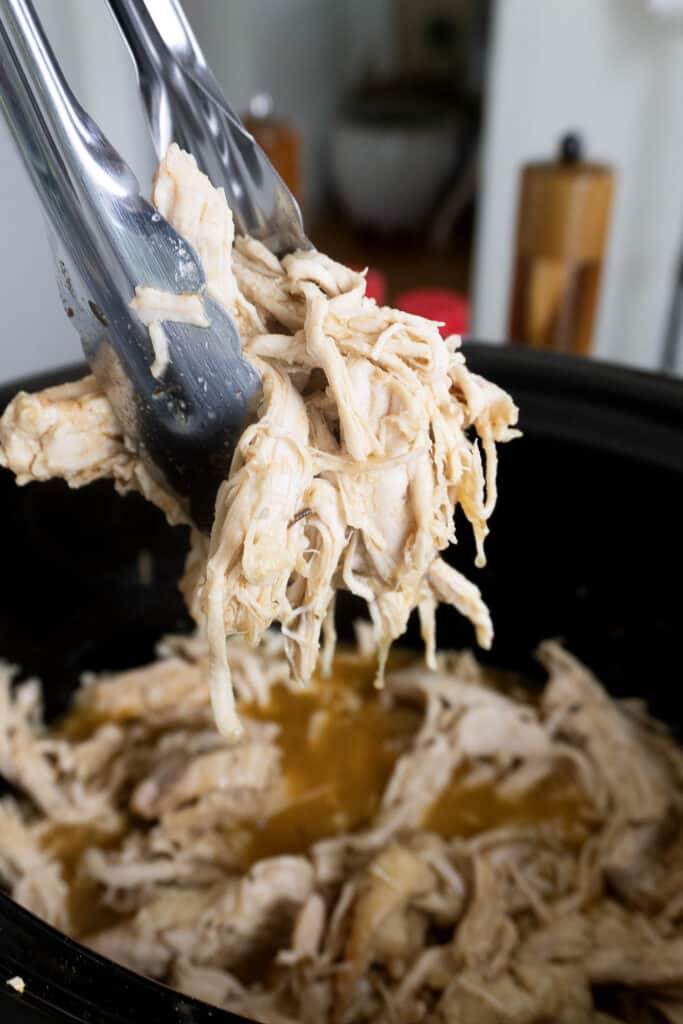 Metal tongs holding shredded chicken over a slow cooker filled with more shredded chicken and broth. The chicken appears moist and tender, ready to be served or used in recipes.