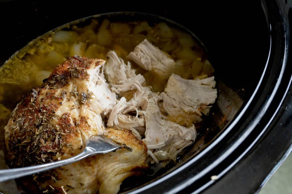 A close-up of cooked, shredded chicken in a slow cooker with broth and herbs, being pulled apart with a fork.