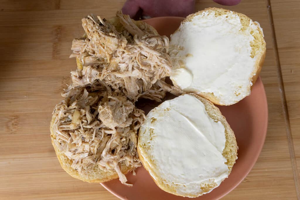 A sandwich bun split open on a plate, with one side spread with mayonnaise and the other topped with shredded cooked chicken. The background is a wooden surface.