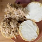 A sandwich bun split open on a plate, with one side spread with mayonnaise and the other topped with shredded cooked chicken. The background is a wooden surface.