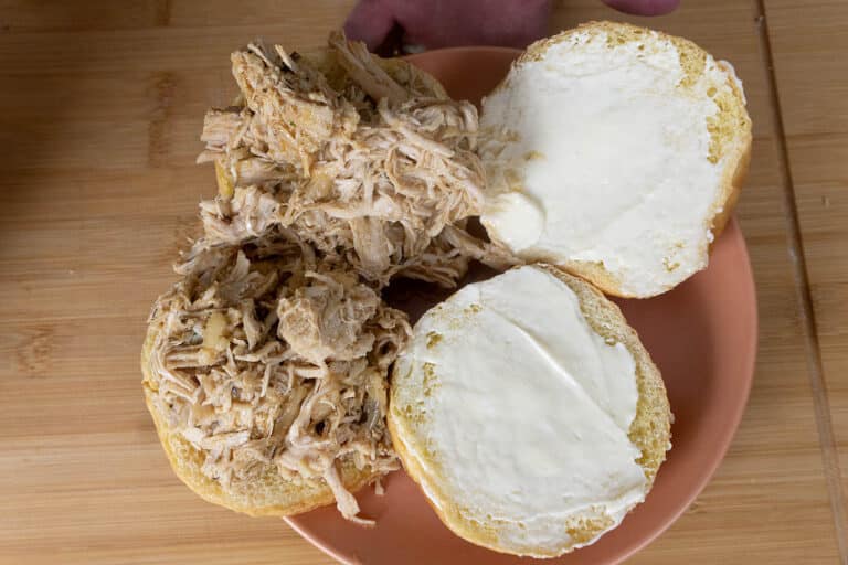 A sandwich bun split open on a plate, with one side spread with mayonnaise and the other topped with shredded cooked chicken. The background is a wooden surface.
