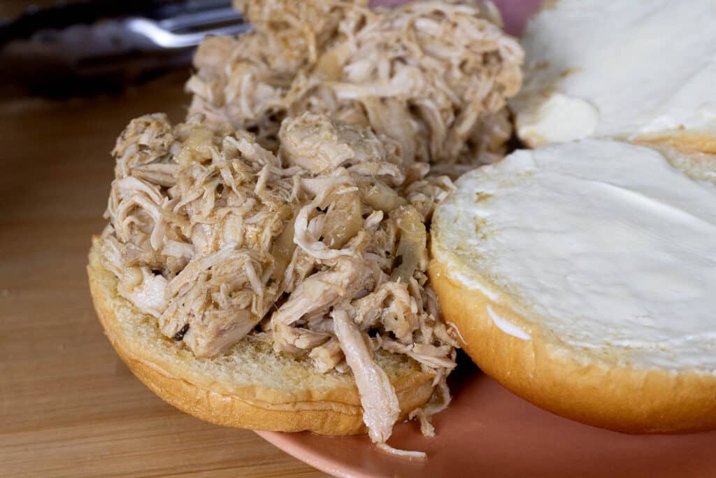 A close-up of a sandwich with shredded chicken piled on one half of a bun and a layer of mayonnaise spread on the other half, all on a pink plate.