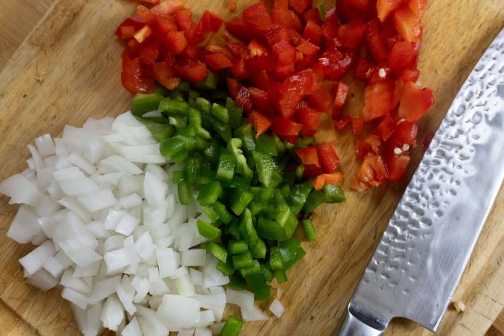 Diced onions, green bell peppers, and red bell peppers on a wooden cutting board next to a large silver knife.