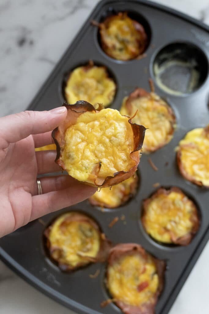 A hand holds a baked egg muffin with cheese in front of a muffin tin filled with similar egg muffins on a marble countertop.