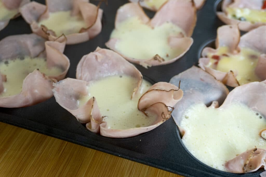 Muffin tin filled with slices of ham shaped into cups and filled with beaten eggs, ready to be baked.