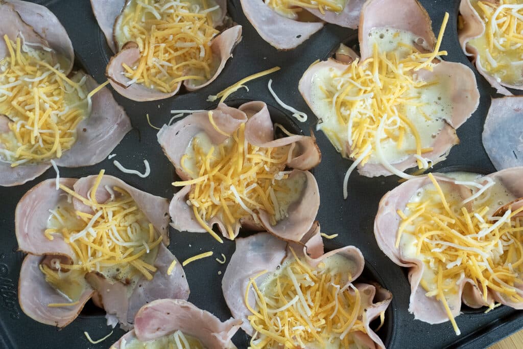 Ham slices in muffin tins, each filled with whisked eggs and topped with shredded cheddar and mozzarella cheese, ready to be baked.