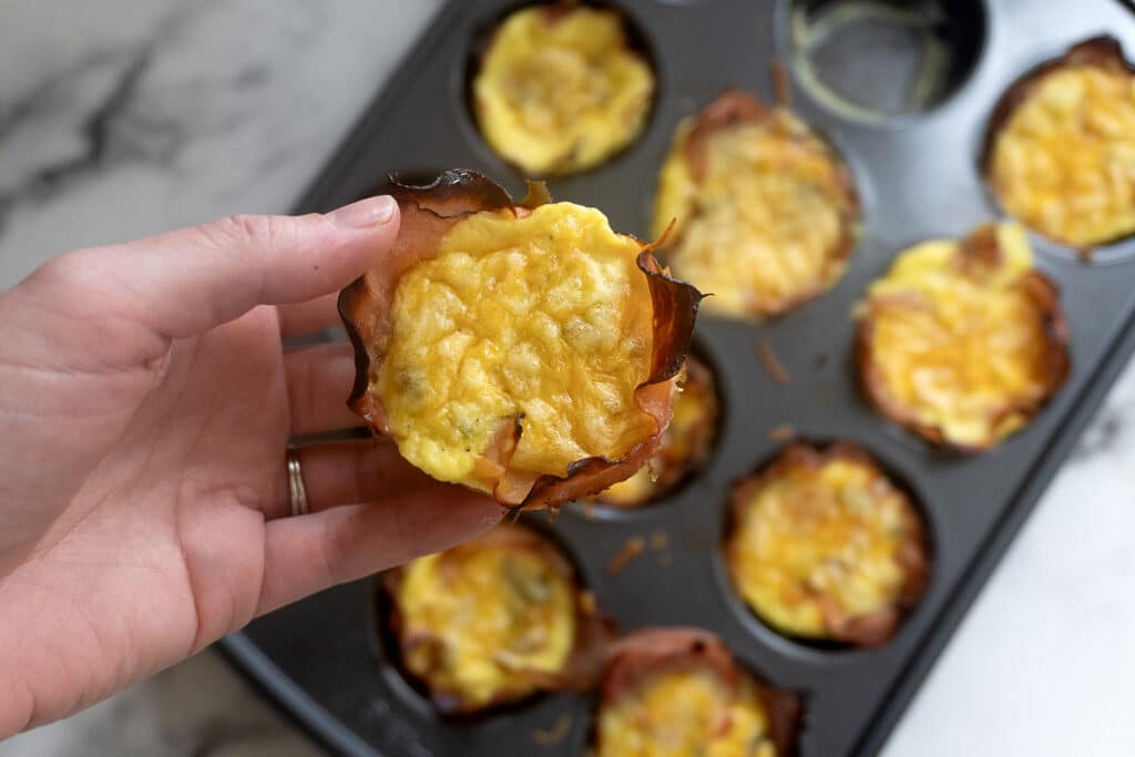 A hand holds a baked egg muffin with a crispy outer layer above a muffin tin filled with similar egg muffins, some missing. The surface beneath is white and marbled.