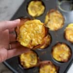 A hand holds a baked egg muffin with a crispy outer layer above a muffin tin filled with similar egg muffins, some missing. The surface beneath is white and marbled.