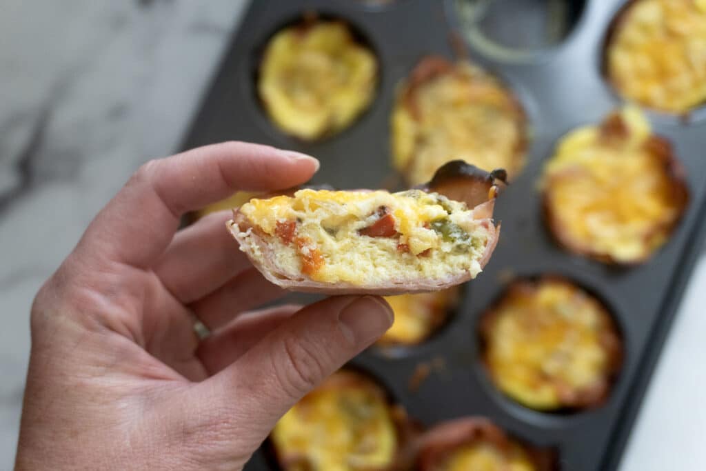 A hand holds a mini quiche with a visible cross-section, showing egg, cheese, and veggies. More quiches are in a muffin tin in the background on a marble surface.