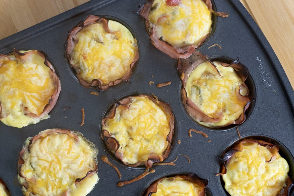 A muffin tin filled with baked egg cups lined with ham and topped with melted cheese, sitting on a wooden surface.