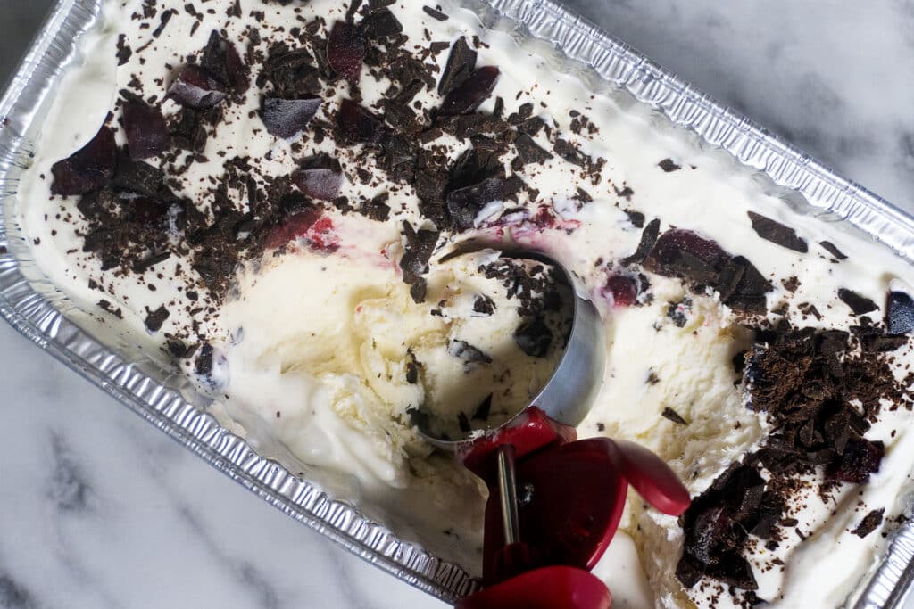 A metal pan of vanilla ice cream topped with chocolate shavings and cherries, with a red-handled ice cream scoop partially embedded, sitting on a marble surface.
