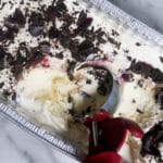 A metal pan of vanilla ice cream topped with chocolate shavings and cherries, with a red-handled ice cream scoop partially embedded, sitting on a marble surface.