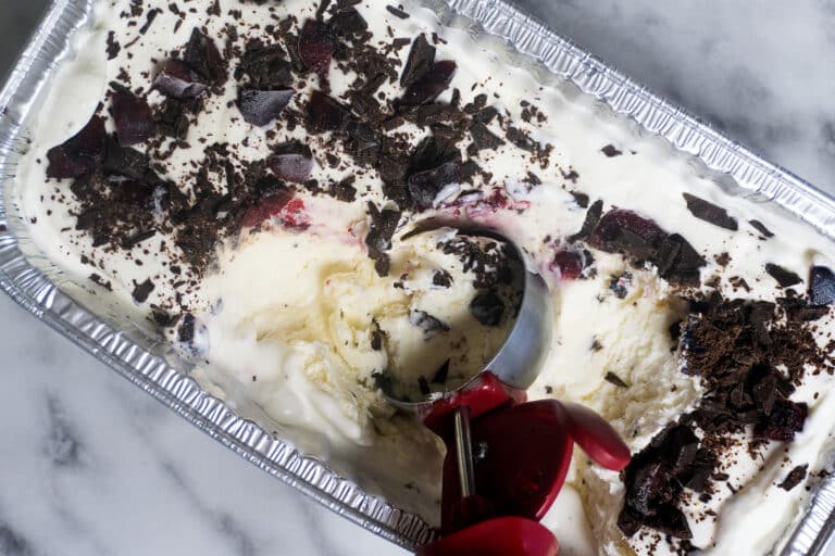 A metal pan of vanilla ice cream topped with chocolate shavings and cherries, with a red-handled ice cream scoop partially embedded, sitting on a marble surface.