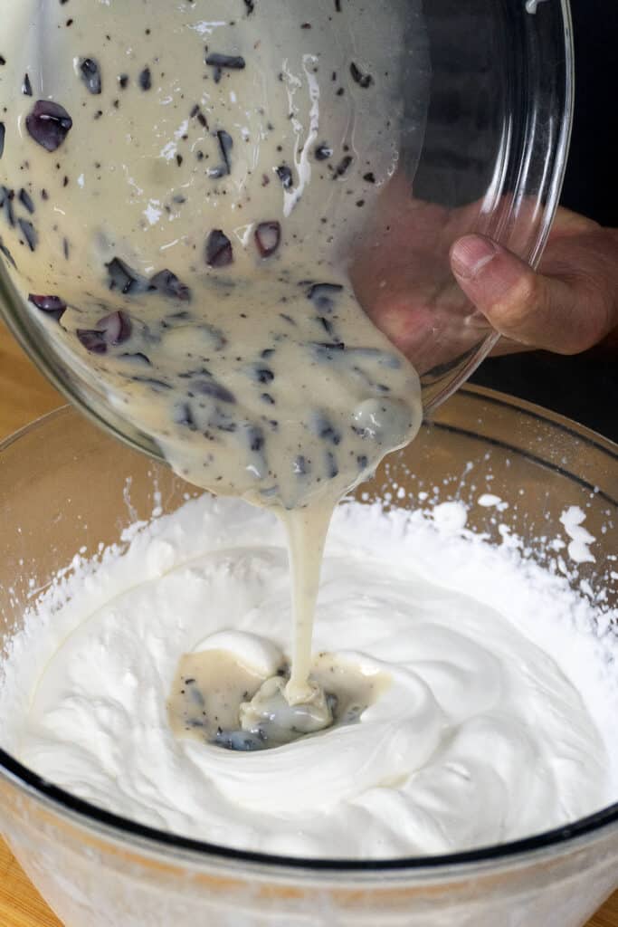 A person pours a thick mixture with dark chunks from a glass bowl into a larger bowl of fluffy white whipped cream on a wooden surface.