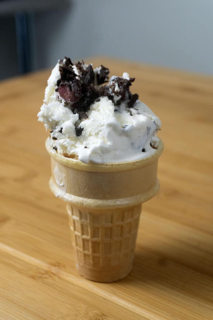 A scoop of vanilla ice cream with chocolate chunks and cherries served in a waffle cone, placed on a wooden table.