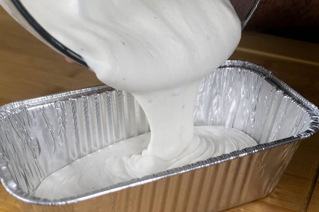 A thick, creamy white mixture is being poured from a glass bowl into a rectangular aluminum foil pan on a wooden surface.