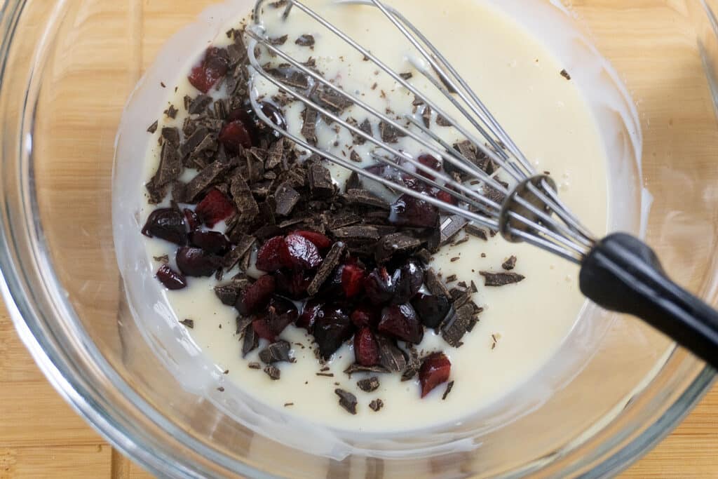 A glass bowl with sweetened condensed milk, chopped dark chocolate, and cherry pieces being mixed with a metal whisk on a wooden surface.