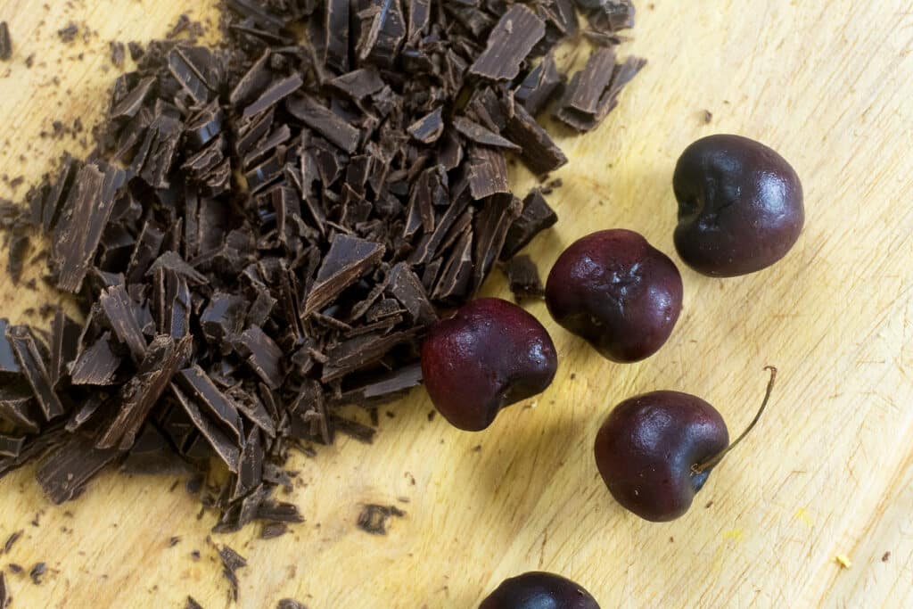 Chopped dark chocolate pieces and five dark red cherries on a wooden cutting board.