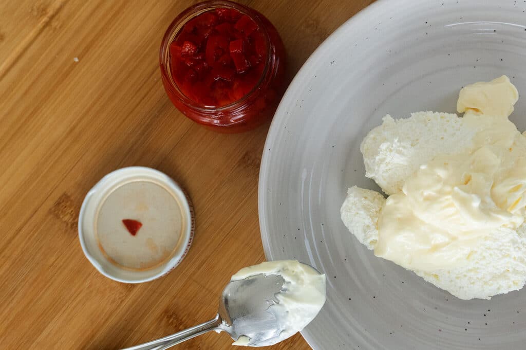 A plate with scoops of white cream or cheese, a spoon with cream, a jar of red preserves, and a lid on a wooden surface.