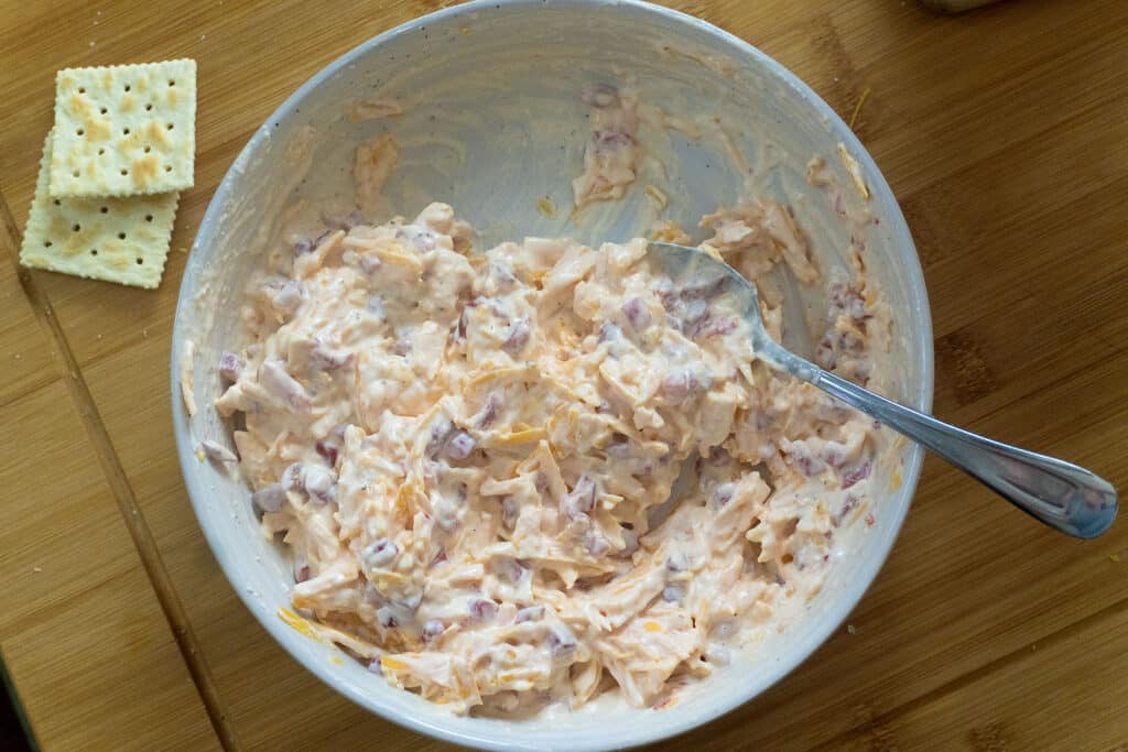 A bowl of creamy pimento cheese spread with a spoon, next to two saltine crackers on a wooden surface.