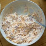 A bowl of creamy pimento cheese spread with a spoon, next to two saltine crackers on a wooden surface.