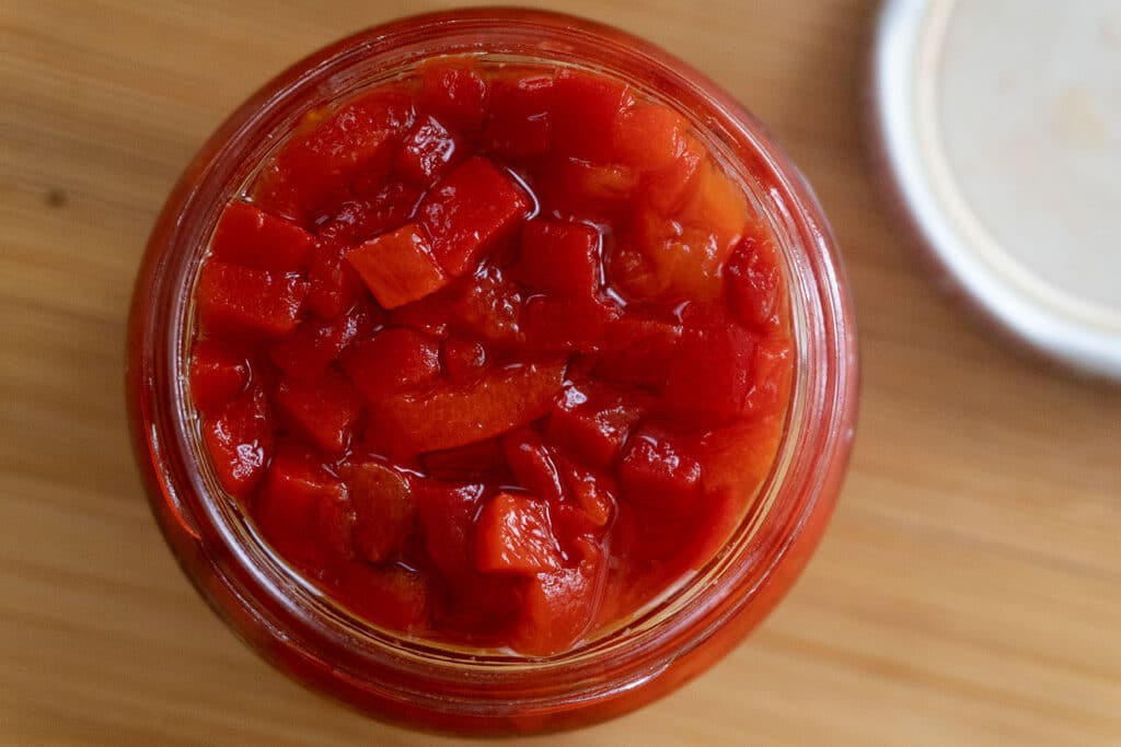 A top-down view of a glass jar filled with chopped red peppers in liquid, sitting on a wooden surface with part of a jar lid visible to the side.