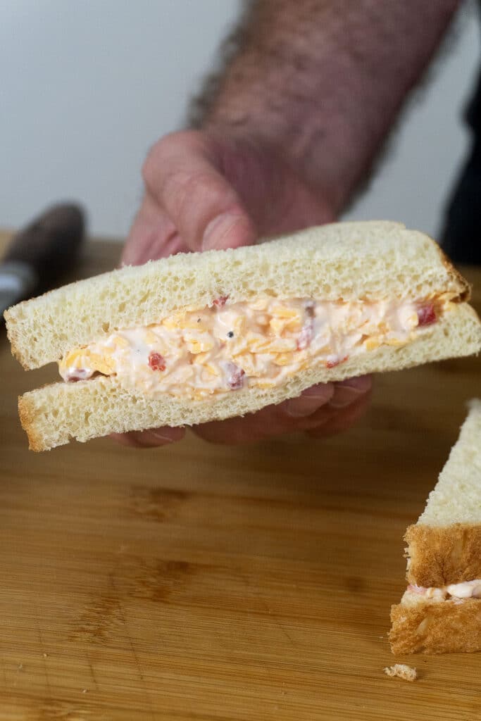 A hand holding a sandwich with creamy pimento cheese filling, made with shredded cheese and diced pimentos, on white bread over a wooden surface.
