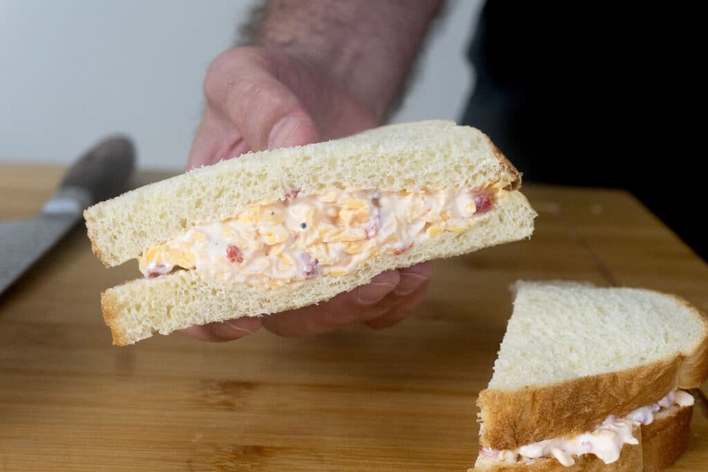 A hand holds a sandwich with a creamy filling, possibly pimento cheese, on white bread. Another sandwich half and a bread knife are on a wooden cutting board in the background.