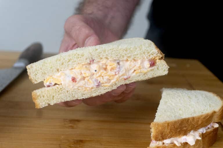 A hand holds a sandwich with a creamy filling, possibly pimento cheese, on white bread. Another sandwich half and a bread knife are on a wooden cutting board in the background.