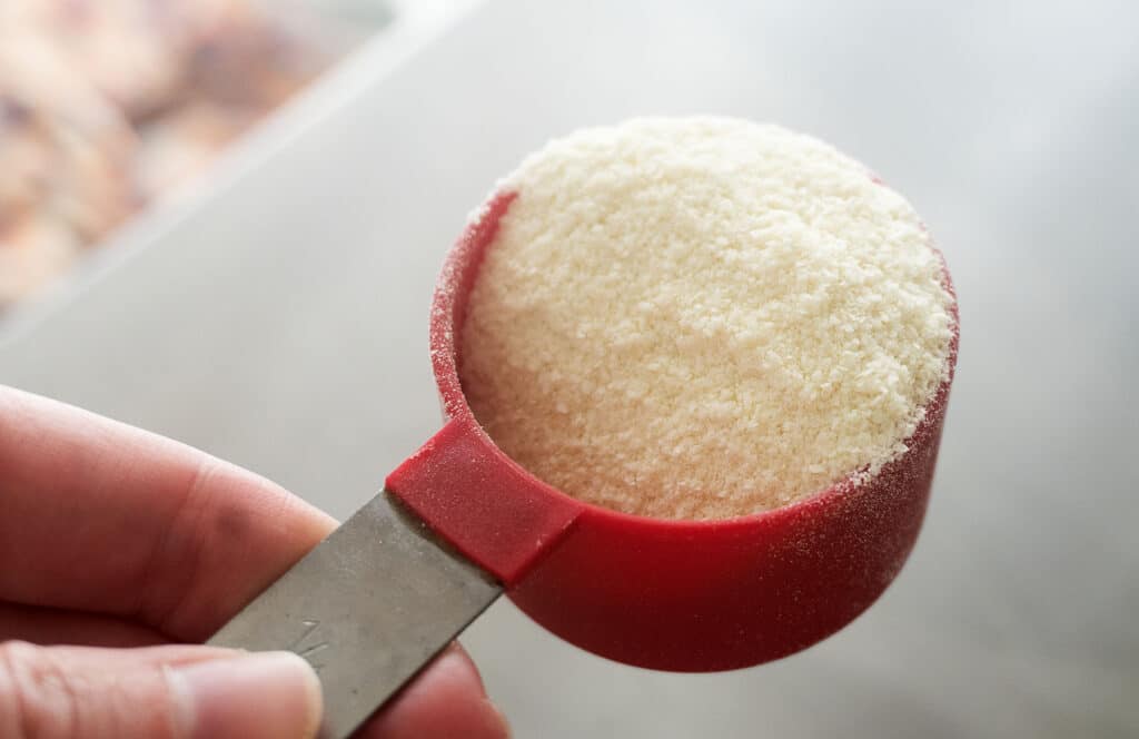 A hand holds a red measuring cup filled with finely grated potato flakes over a gray surface.